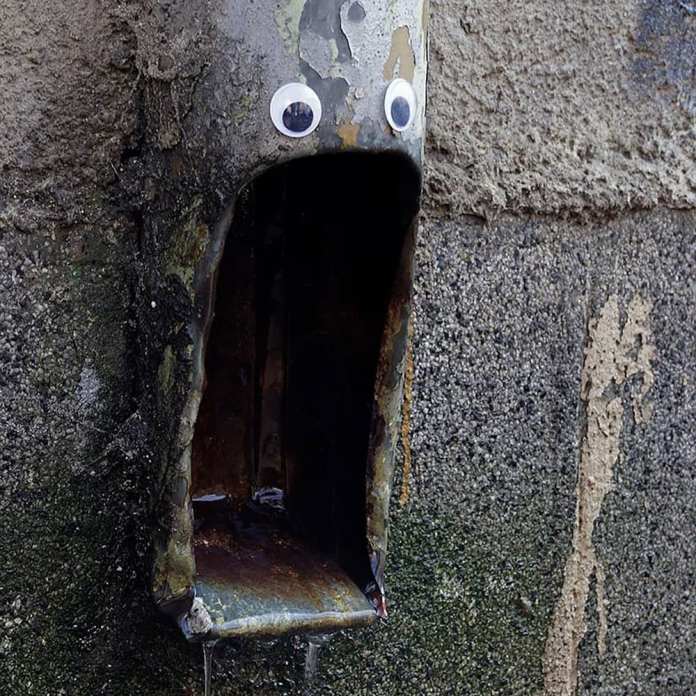 A quirky piece of googly eye street art in Bulgaria, featuring a rusted metal drainage pipe attached to a textured concrete wall. The pipe, with its dark interior resembling an open mouth, is brought to life with two googly eyes placed above it, creating the impression of a shocked or surprised expression. The surrounding wall shows signs of weathering, adding to the raw, urban feel of this playful and creative intervention.