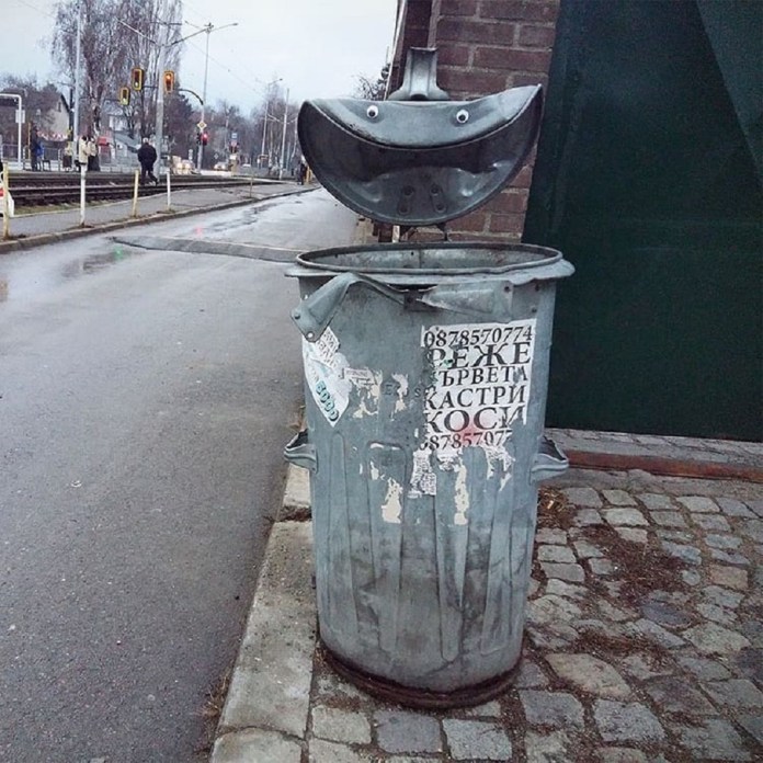 A humorous piece of googly eye street art in Bulgaria, where a metal trash can has been transformed into a cheerful character. The open lid of the trash can resembles a wide, smiling mouth, and two googly eyes are placed on the top, giving it a friendly, animated appearance. The scene is set on a rainy urban street with tram tracks, traffic lights, and pedestrians in the background, emphasizing the integration of this playful art into everyday city life. The trash can also features worn-out stickers and text, adding to the raw and authentic urban vibe.
