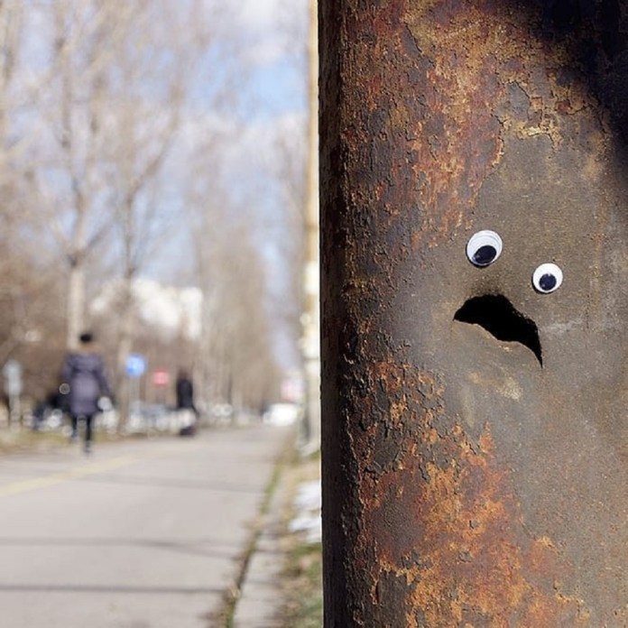 A creative and humorous example of googly eye street art in Bulgaria, featuring a rusty metal pole with two googly eyes and a small crack resembling a surprised or worried mouth. The simple addition of the eyes transforms the pole into a character full of emotion. The background shows a tree-lined sidewalk with pedestrians, adding an urban context to this playful and imaginative street art piece. The rust and texture of the pole highlight the raw, unpolished charm of this urban intervention.