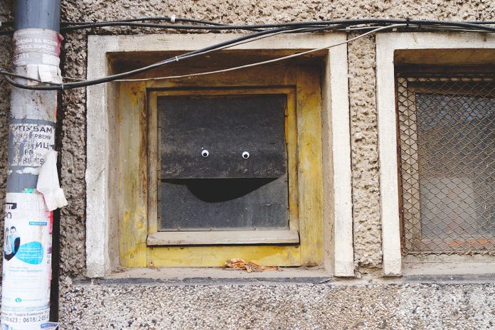 A playful piece of googly eye street art in Bulgaria, featuring a boarded-up window on a textured wall. The window has been transformed into a cheerful character by adding two googly eyes on the upper board and creating a smile with the curved edge of the lower board. The surrounding wall is rough and urban, with visible electrical wires and a pole covered in torn posters, adding to the gritty city setting. This creative intervention brings humor and personality to an otherwise ordinary and overlooked urban element.