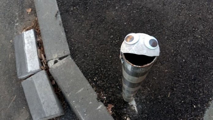 A humorous piece of googly eye street art in Bulgaria, featuring a broken metal post transformed into a cheerful character. The post, with its jagged open top resembling a mouth, has two large googly eyes placed on the rim, giving it a happy and animated expression. It is situated on a paved street corner with asphalt and concrete curbs surrounding it. This creative intervention adds a playful and unexpected touch to an otherwise mundane urban element.
