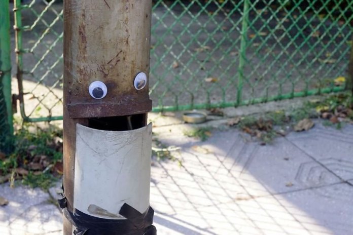 A fun piece of googly eye street art in Bulgaria, featuring a rusty metal pole turned into a character with a face. Two googly eyes are attached to the upper part of the pole, and a cutout on the lower part resembles a slightly open mouth. The pole is set against a green wire fence and a paved sidewalk, with fallen leaves and shadows adding to the urban outdoor setting. This creative and whimsical transformation brings a sense of humor and personality to an ordinary street element.