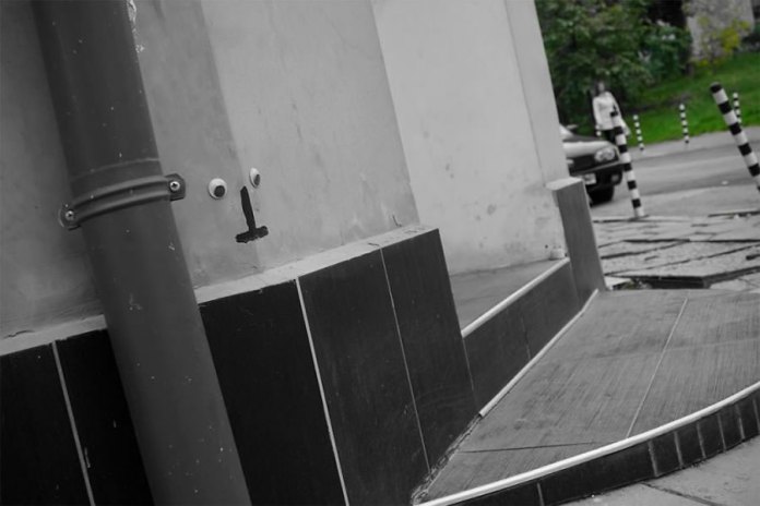 A minimalist piece of googly eye street art in Bulgaria, where a small face is created on a building wall. Two googly eyes are attached near a pipe, with a simple black line painted below to represent a nose or mouth, giving the impression of a quirky character peeking from the corner. The scene includes a tiled ramp, a parked car, and a blurred figure in the background, emphasizing the urban environment. The grayscale tones with selective green in the background enhance the artistic and playful feel of this intervention.