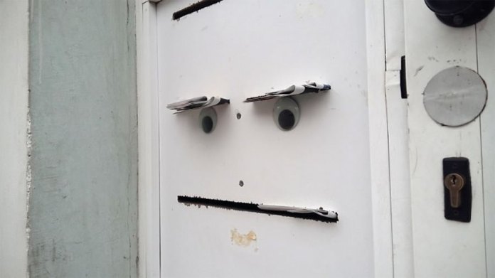 A clever piece of googly eye street art in Bulgaria, featuring a white door transformed into a face. Two mail slots with googly eyes attached beneath them resemble heavy-lidded eyes, while a small slit below forms a straight mouth, creating a grumpy or unimpressed expression. The minimalistic design integrates everyday elements into a humorous and relatable character, bringing personality to a mundane setting.