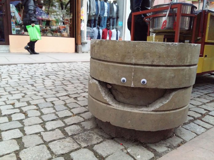 A playful piece of googly eye street art in Bulgaria, featuring a cracked concrete barrier turned into a smiling character. The crack resembles a wide, open mouth, and two googly eyes are placed above it, creating a cheerful expression. The barrier sits on a cobblestone street, with a storefront displaying clothing and a street vendor's cart in the background. This creative and humorous transformation adds a sense of whimsy to the bustling urban environment.