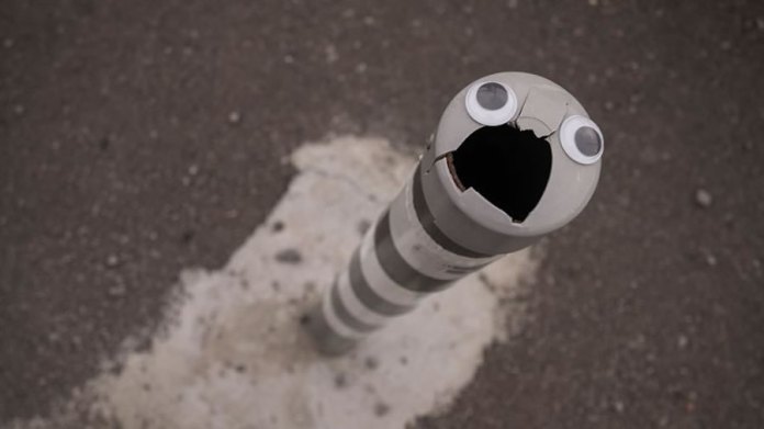 A humorous piece of googly eye street art in Bulgaria, featuring a broken bollard with a striped pattern. The jagged top of the bollard resembles an open mouth, and two googly eyes are placed above it, creating an expressive, surprised face. The bollard is surrounded by asphalt and a patch of rough concrete, emphasizing the urban setting. This creative and simple transformation brings personality and humor to a mundane street element.