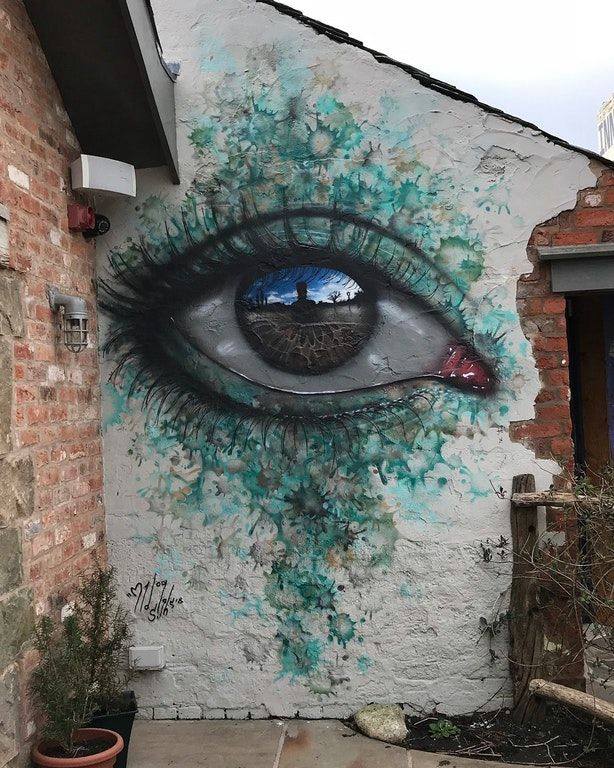 Large eye mural painted on the exterior of a brick and stucco wall by artist My Dog Sighs in Eccleston, England. The mural features a strikingly realistic eye with intricate eyelashes and shading, surrounded by a textured green-blue splash effect that resembles water or paint splatter. The eye reflects a scene of a person standing in front of a stone pathway and open sky, adding depth to the artwork. The signature 'My Dog Sighs' is visible near the lower left of the piece, blending naturally with the weathered wall textures and small plants around the base.