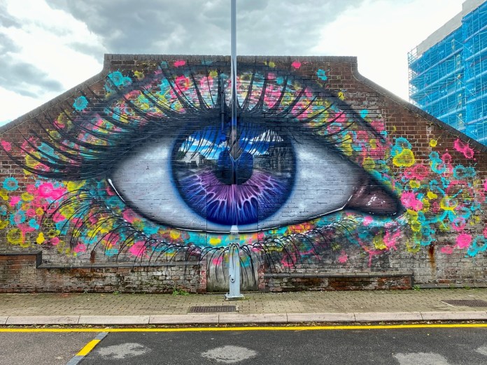 Massive eye mural by artist My Dog Sighs on a brick building wall in Portsmouth, UK, created for LOOK UP Portsmouth. The mural features a hyper-realistic blue and purple iris with a reflective surface, showing faint images of nearby buildings and a lamppost, adding depth to the artwork. The eye is surrounded by colorful splashes and streaks of neon pink, yellow, and blue paint, giving it a vibrant, urban aesthetic. The fine details in the eyelashes and shading add to the surreal effect, making the eye appear almost alive against the worn brick background. A lamppost stands directly in front of the mural, blending into the artwork and further enhancing the illusion of depth.