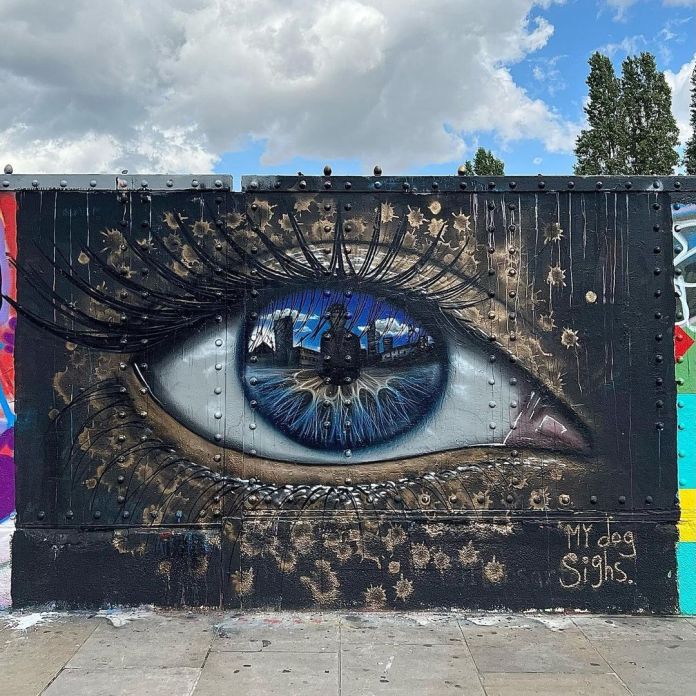 Eye mural by artist My Dog Sighs on a textured metal wall in London, UK, photographed by Brian B. The mural depicts a hyper-realistic blue eye with intricate details, including reflections of buildings and a cloudy sky, giving the iris a glassy, lifelike quality. The wall's metallic surface, complete with rivets and industrial wear, adds a gritty backdrop to the artwork, making the eye appear as though it’s embedded in metal. Surrounding the eye are rust-colored splatters and scratches, enhancing the rugged and worn aesthetic. The artist's signature, 'My Dog Sighs,' is visible in the bottom right corner, blending with the wall’s urban textures.
