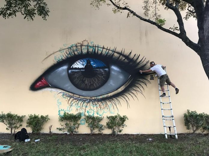 Eye mural in progress by artist My Dog Sighs on a wall at Santa Clara Elementary School in Wynwood, Miami, Florida, part of the aWall Mural Projects. The mural showcases a hyper-realistic eye with a reflective iris that mirrors the image of the artist and nearby surroundings, adding a sense of depth and presence. The eye has intricate detailing, with delicate eyelashes and a tear duct painted in red. Surrounding the eye are subtle aqua-colored paint splatters, enhancing the piece’s vibrant, surreal quality. The artist is seen on a ladder, adding final touches to the mural, demonstrating the scale and detail of the artwork. Small bushes and green grass line the bottom of the wall, framing the mural against the beige surface.