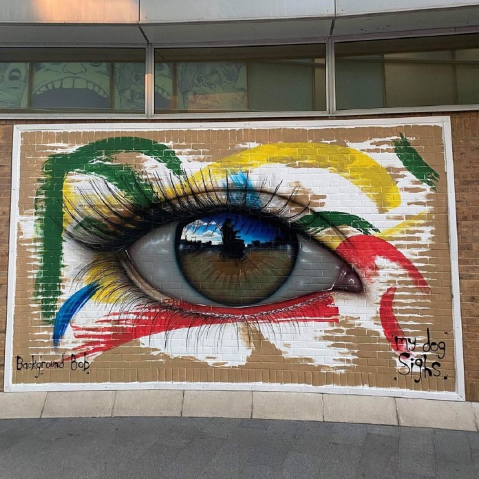 Eye mural collaboration by artists My Dog Sighs and Background Bob on a brick wall in Southampton, UK. The mural depicts a hyper-realistic eye with a brown iris, reflecting the skyline of a city with clouds in the sky, adding depth to the artwork. The eye is surrounded by vibrant brushstrokes in red, yellow, green, and blue, creating an abstract background that contrasts with the realistic detail of the eye. The artists’ signatures, 'My Dog Sighs' and 'Background Bob,' are visible near the lower edge, acknowledging their collaboration. The artwork is framed within a brick wall section, with a building and windows visible above, blending urban surroundings with the mural's striking imagery.