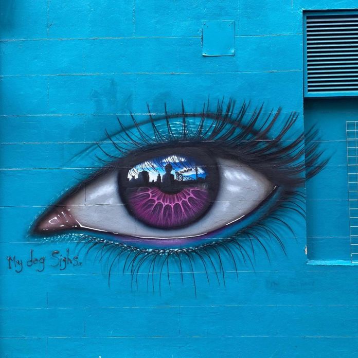 Eye mural by artist My Dog Sighs on a vibrant blue wall in Glasgow, Scotland. The mural features a striking eye with a deep purple iris, displaying intricate details and a reflective quality that mirrors the cityscape, including buildings and a partly cloudy sky. The eyelashes are long and detailed, adding to the realistic effect, while the pinkish-red tear duct contrasts with the bold blue background. The artist’s signature, 'My Dog Sighs,' is visible near the lower left corner, blending into the wall’s smooth surface. This mural, set against the bright blue wall, creates a powerful focal point with its surreal and expressive detail.