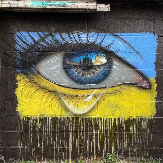 Emotive eye mural by artist My Dog Sighs on a wall in Cardiff, Wales, symbolizing solidarity with Ukraine amidst the Russian invasion. The mural features a hyper-realistic blue eye with a reflection of Kyiv's skyline engulfed in flames and smoke, representing the turmoil of the invasion. The background of the mural incorporates the colors of the Ukrainian flag, with blue on top and yellow below, both slightly textured and dripping to add a raw, distressed effect. A single tear falls from the eye, blending into the yellow section and symbolizing sorrow and resilience. The artwork powerfully conveys the artist's sadness and anger over the conflict, using the eye as a poignant metaphor for watching the tragic events unfold.