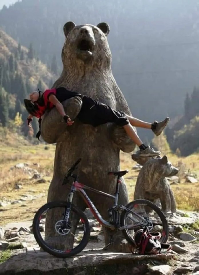 A cyclist in red gear poses limp in the arms of a large bear statue with two cubs nearby, creating a humorous wildlife rescue scene in a mountainous landscape.