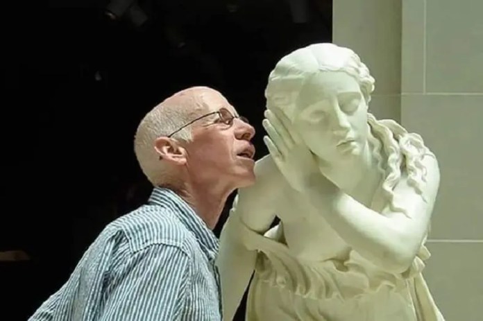 A bald man whispers into the ear of a white marble statue of a woman who appears to be listening, set indoors against a dark background.