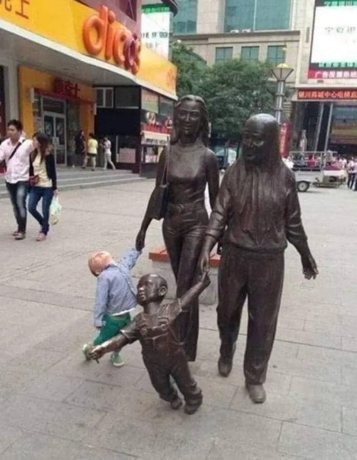 A toddler in green pants and blue shirt walks alongside a bronze statue group, unintentionally matching the forward-reaching pose of the sculpted child in the scene.