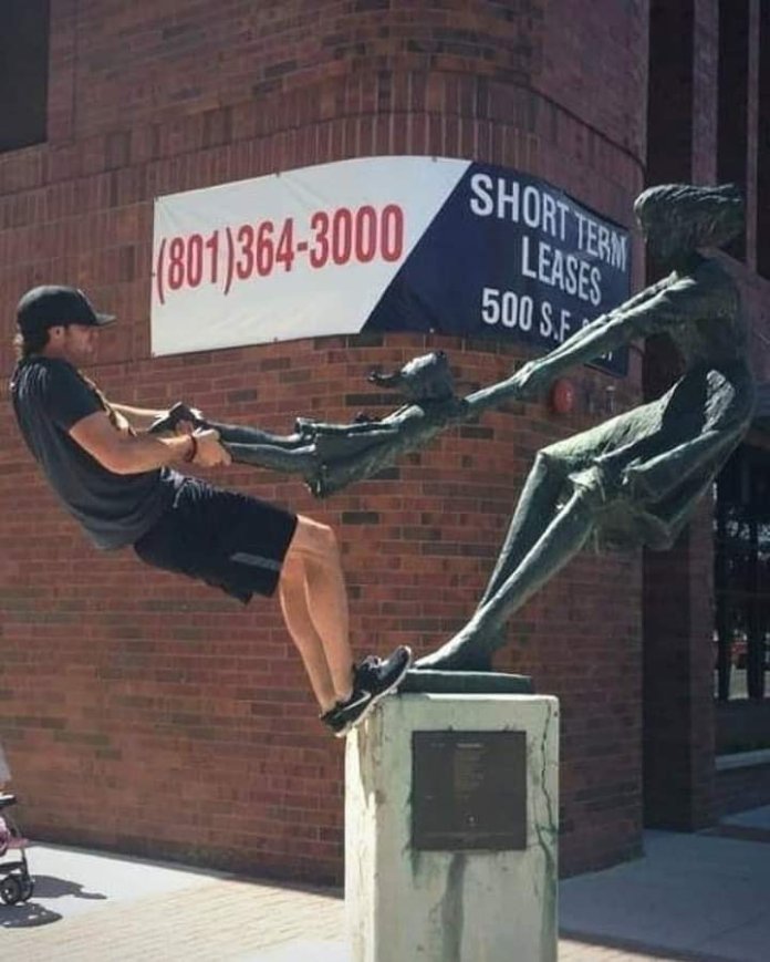 A man leans back in an exaggerated tug-of-war with a bronze sculpture called "The Traveler" in Salt Lake City, Utah. This piece, which shows a figure extending their hand forward, is a popular attraction for playful photos, as it invites people to interact with its dynamic pose.