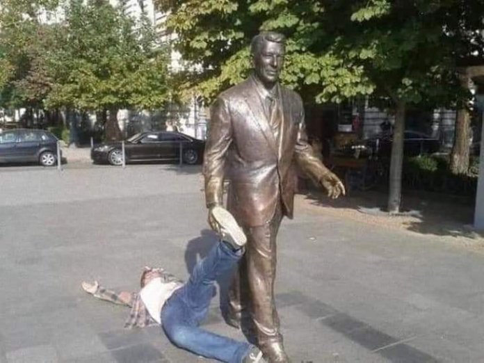 A person humorously "falls" at the foot of the Ronald Reagan statue in Budapest, Hungary, adding a lighthearted moment to the iconic sculpture.