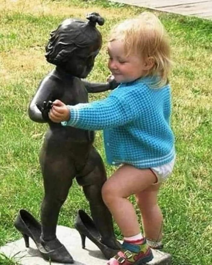 A young child holds hands with a statue of a small girl wearing oversized shoes. This charming moment captures the innocence and imagination that art can inspire, as the child seems to engage in a make-believe friendship with the statue.