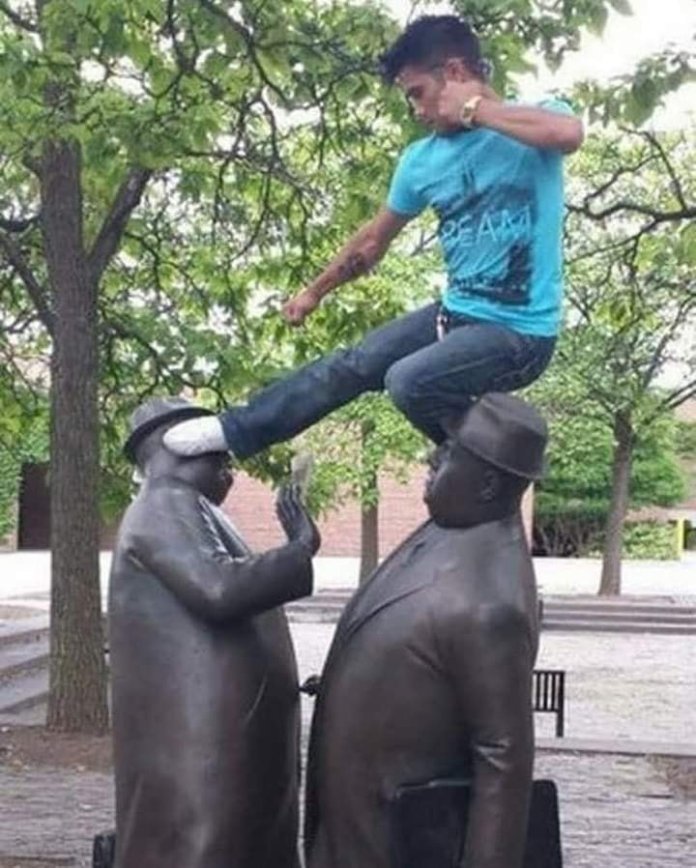 This image shows a person playfully pretending to kick between two statues mid-conversation, bringing action and modern humor to the scene. The playful energy contrasts with the serious demeanor of the statues.