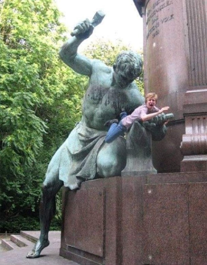 A man humorously interacts with a large statue of a muscular figure wielding a hammer and chisel, as if the statue is about to strike him. The man leans against the statue, looking surprised, adding a playful element to the powerful pose of the sculpture. The scene takes place outdoors, with greenery visible in the background. The statue itself is mounted on a stone pedestal, creating an imposing yet amusing interaction between art and visitor.