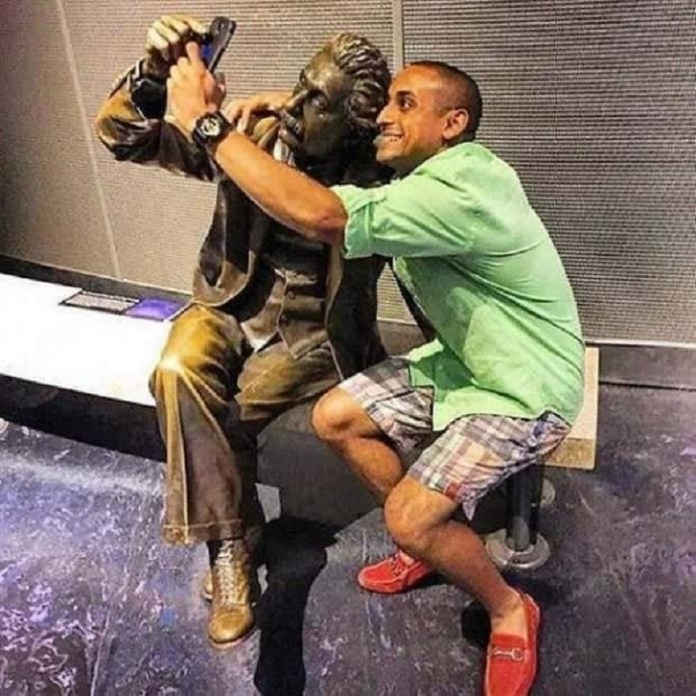 A man takes a selfie with a bronze statue of Albert Einstein, playfully positioning the phone as if Einstein himself is posing for the photo. The man, dressed in a green shirt, plaid shorts, and red loafers, grins as he leans in close to the statue in a friendly, humorous moment. The setting appears to be an indoor exhibit space, adding a casual and interactive twist to the iconic image of Einstein.