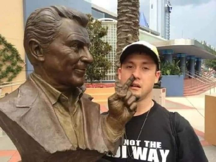 A humorous interaction between a man and a bronze bust statue. The man stands beside the statue, positioning himself so that the statue’s extended finger appears to be poking him in the nose. The playful pose creates a comical moment, with the statue’s friendly expression adding to the lighthearted scene. A modern building and palm trees provide a vibrant background.