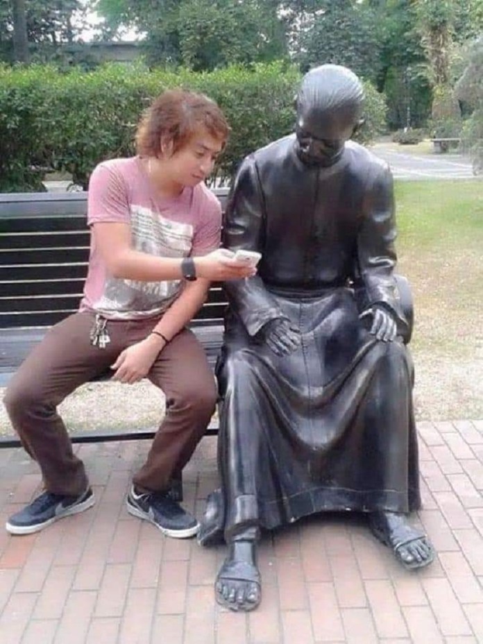 A young man sitting on a park bench shares something on his phone with a life-sized bronze statue of an elderly figure, creating a humorous moment of connection. The statue, dressed in traditional attire with hands resting on its lap, appears to gaze down at the phone in curiosity. The greenery in the background adds to the serene park setting.