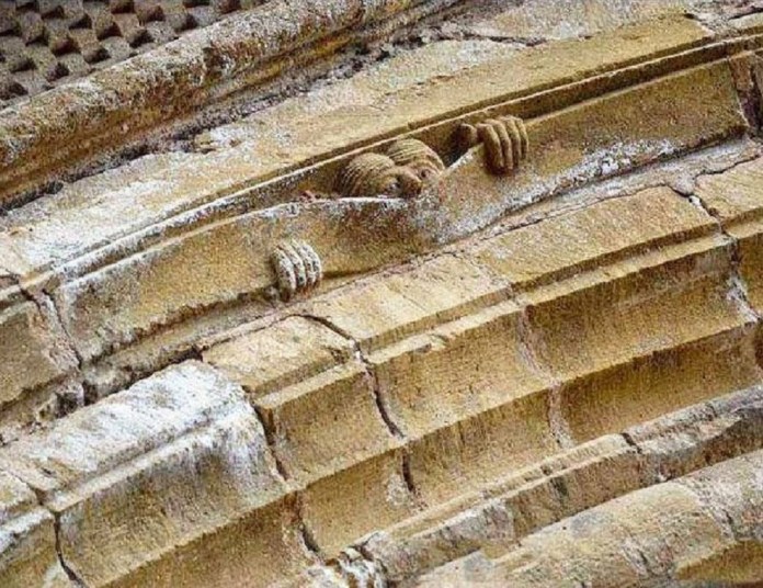 A unique gargoyle sculpture known as 'The Peeker of Conques' at the Abbey of Sainte-Foy in Conques, France, dating from around 1107. The sculpture shows a figure with its hands gripping the edge of the stone, peeking out from the structure. This playful and humorous gargoyle is an example of medieval artisans' creativity, blending function as a water spout with whimsical, fantastical expression.