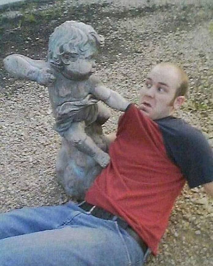 Man in red shirt on the ground with a surprised face as a cherub statue extends an arm toward his chest in an aggressive pose.