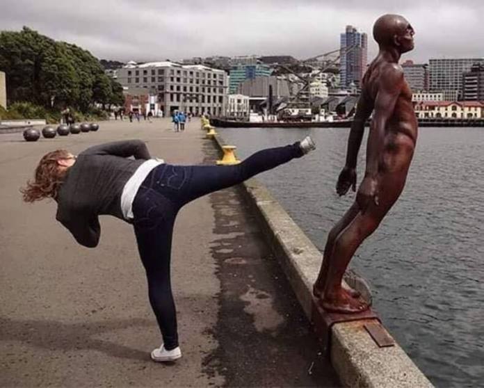 Woman poses with leg extended as if kicking a bronze male statue near the harbor, with city buildings in the background.