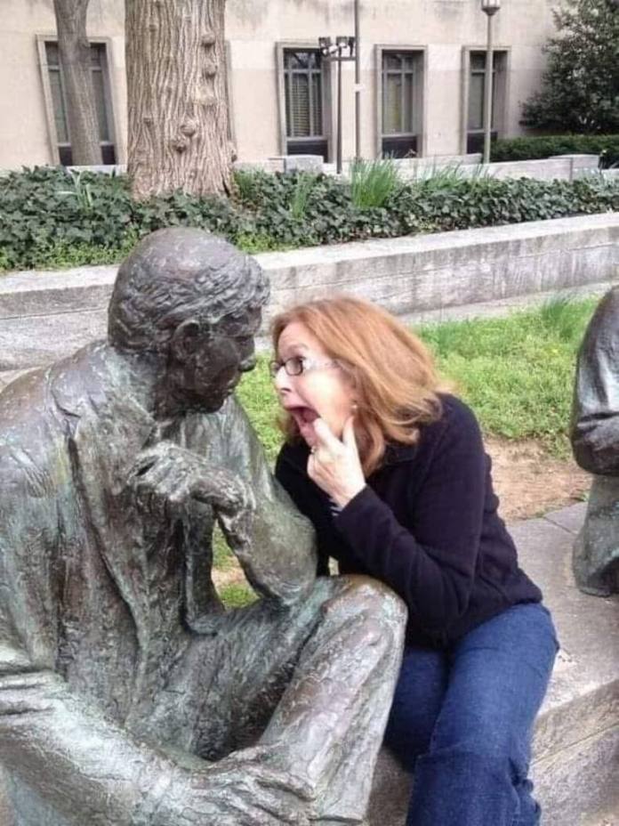 Woman holds her chin and opens her mouth dramatically while facing a bronze statue seated in thought.
