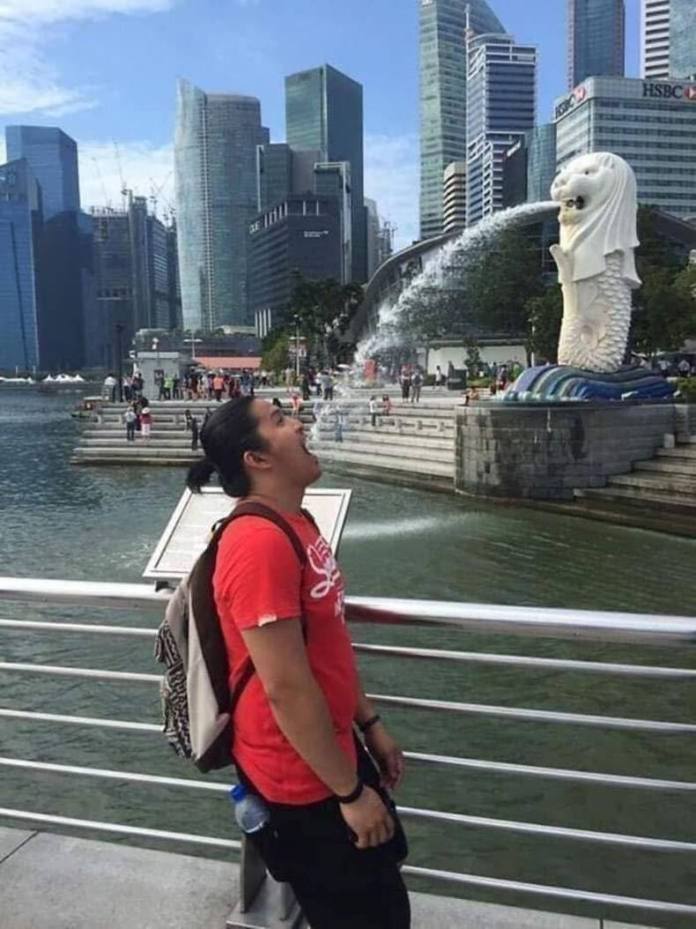 Man in red shirt leans back with mouth open, perfectly aligned with the water spouting from Singapore's Merlion statue.