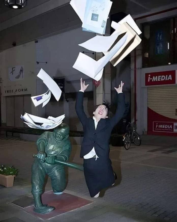 Man in business attire jumps and tosses papers as a nearby bronze statue of a batter swings a bat, suggesting a powerful hit.