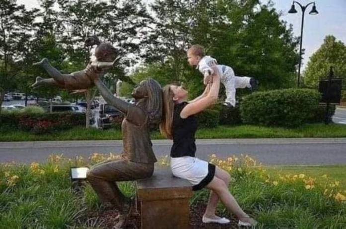 Woman in black and white outfit holds her baby aloft while next to a seated statue lifting a sculpted child in the same pose.