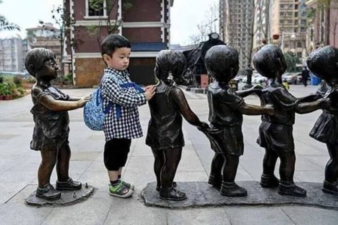 Young boy wearing a checkered shirt and backpack stands between bronze child statues in a line, appearing as part of the sculpture.