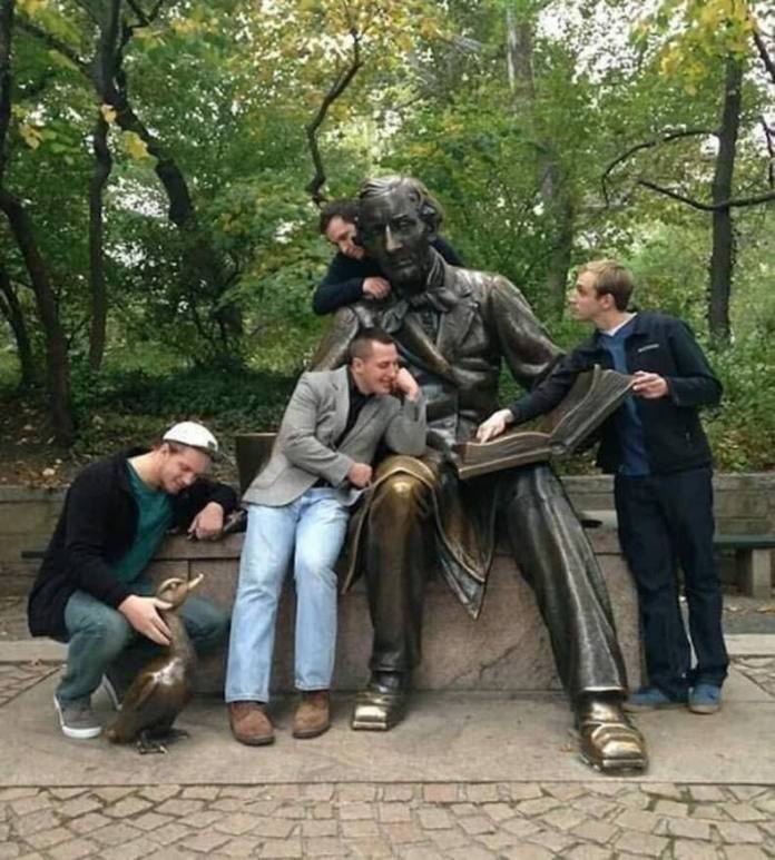 Four men pose around a statue of Hans Christian Andersen reading a book, mimicking listening and reading positions in a park setting.