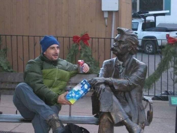 Man in a puffy green jacket and blue hat shares a soda with a statue of a man sitting cross-legged on a bench.
