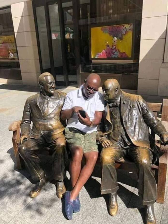 Man in white shirt and shorts sits between two bronze statues on a bench, holding a phone they all appear to be looking at.
