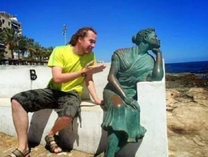 Man gestures and leans toward a green patina statue on a white bench that appears to be turning away in disinterest.