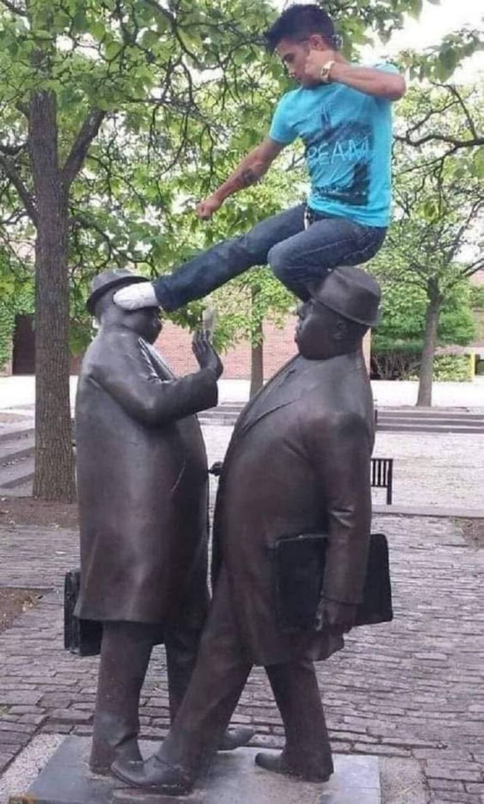 Young man balances between two bronze businessmen statues, delivering a mock martial arts kick to one’s face.