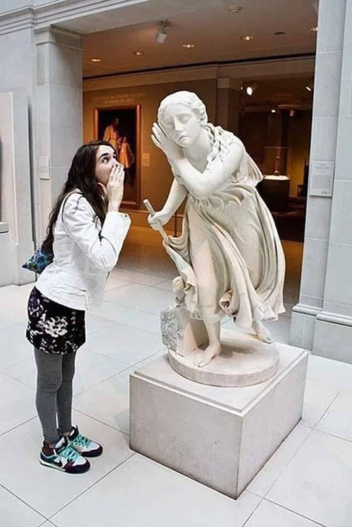 Woman leans forward and whispers into the ear of a marble statue posed with one hand to its ear and the other holding a rod.