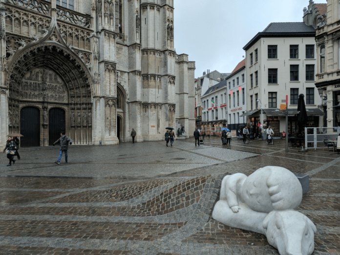 The sculpture of Nello and Patrasche from A Dog of Flanders, created by artist Batist Vermeulen (Tist), lying under a cobblestone blanket near the Cathedral of Our Lady in Antwerp, Belgium. The gothic cathedral's grand facade dominates the background, while the wet cobblestone square adds a reflective atmosphere, highlighting the deep emotional connection depicted in the artwork.