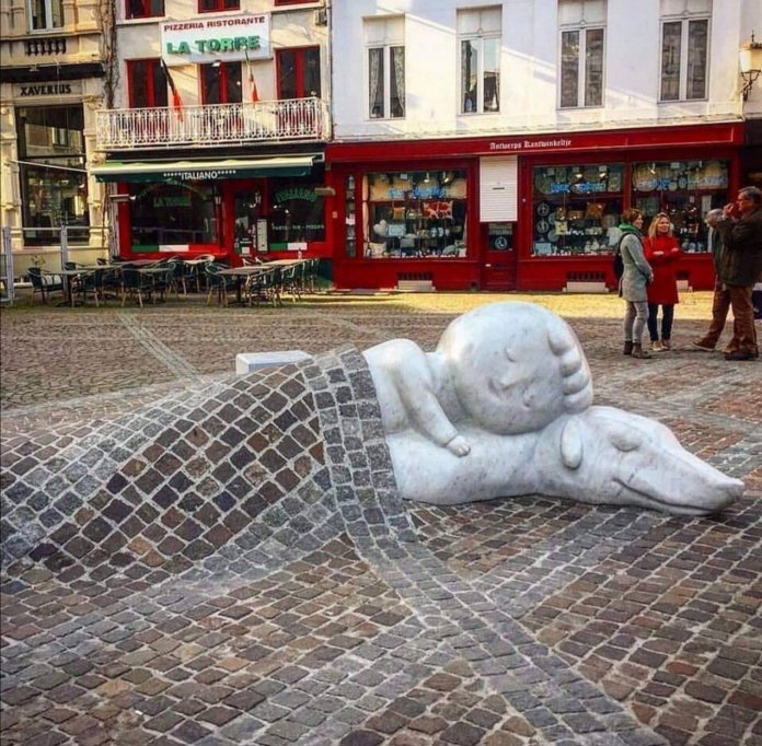 A touching sculpture of Nello and Patrasche, characters from the novel A Dog of Flanders, lying together under a cobblestone blanket. The artwork by Batist Vermeulen, also known as Tist, is located in Antwerp, Belgium, near the Cathedral of Our Lady. The sculpture symbolizes enduring friendship and resilience, set against a vibrant urban backdrop with historic architecture