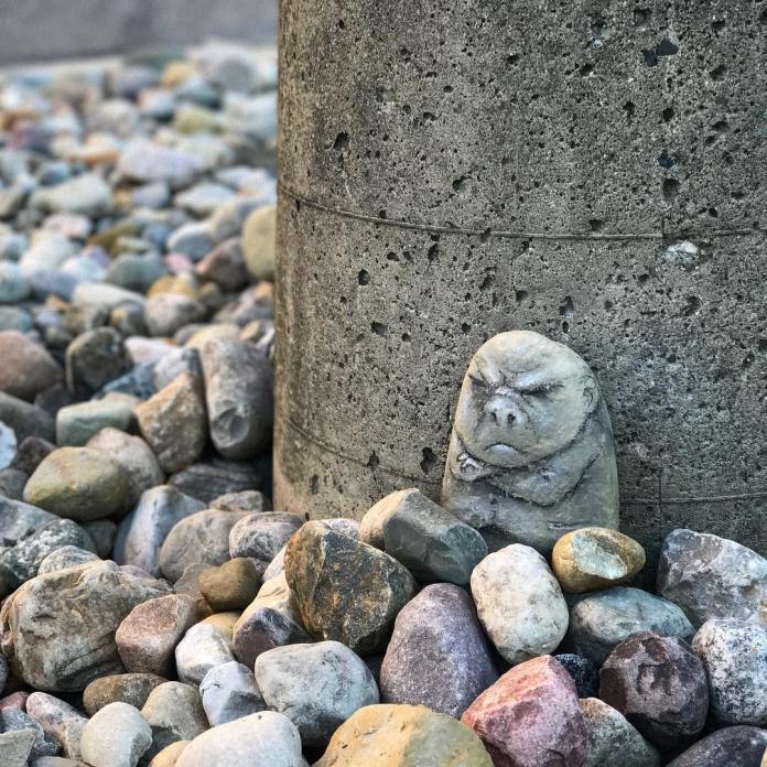 A small stone painted to resemble a grumpy, cartoonish creature with crossed arms. It is placed among other natural rocks at the base of a concrete pillar, almost camouflaged with its surroundings.