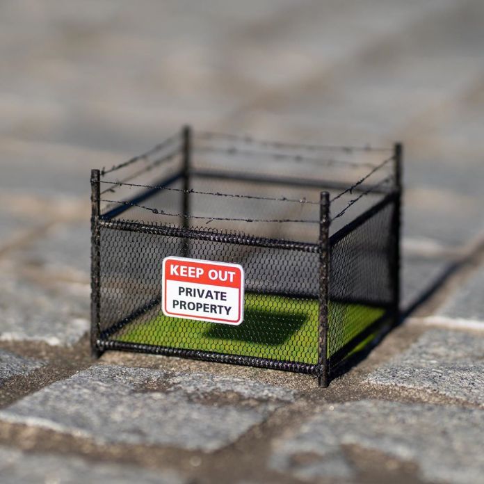 A small patch of artificial grass enclosed by a miniature barbed wire fence with a sign reading "KEEP OUT: PRIVATE PROPERTY." This humorous street art piece by Michael Pederson in Sydney, Australia, critiques the concept of exclusivity in public spaces.