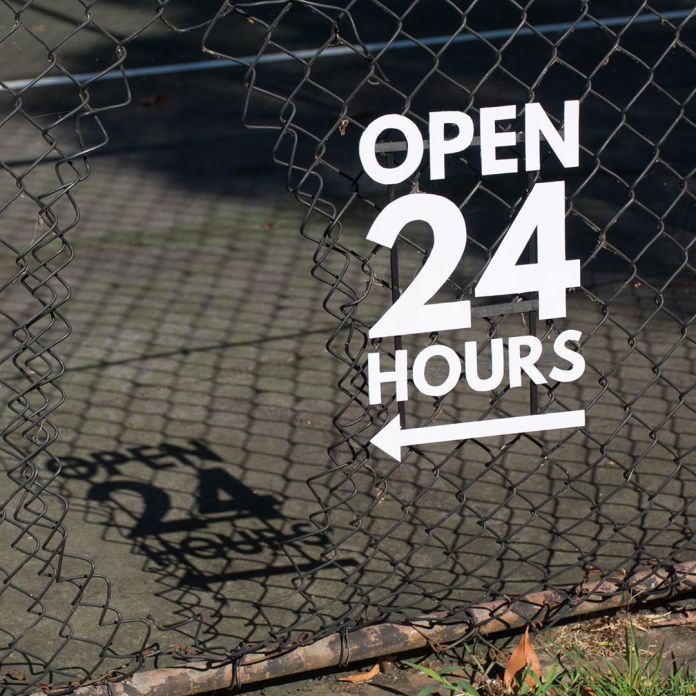 A damaged chain-link fence with a white sign reading "OPEN 24 HOURS" and an arrow pointing to the right. The shadow of the text is cast on the ground, humorously contrasting the dilapidated state of the fence with the suggestion of accessibility. This is a street art installation by Michael Pederson in Sydney, Australia.