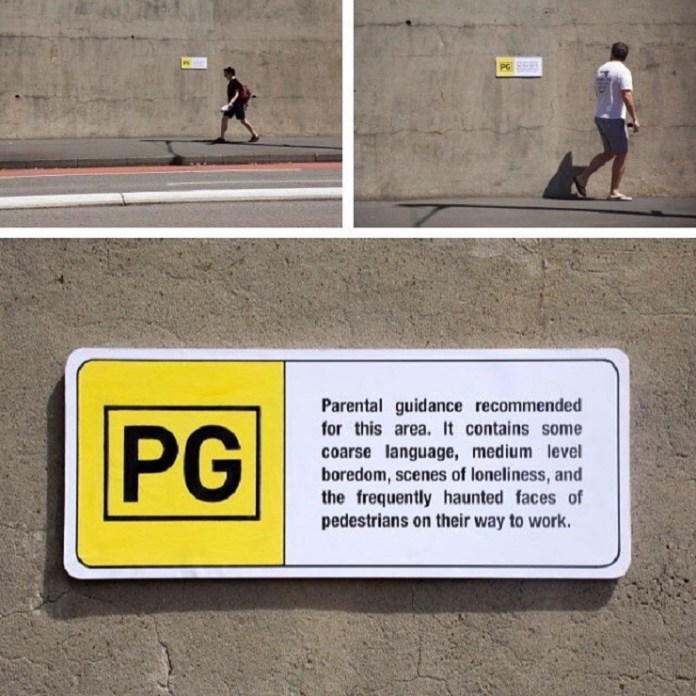 A mock "PG" rating sign installed on a plain concrete wall by Michael Pederson in Sydney, Australia. The sign humorously reads: "Parental guidance recommended for this area. It contains some coarse language, medium level boredom, scenes of loneliness, and the frequently haunted faces of pedestrians on their way to work." The artwork playfully critiques the monotony of urban environments.