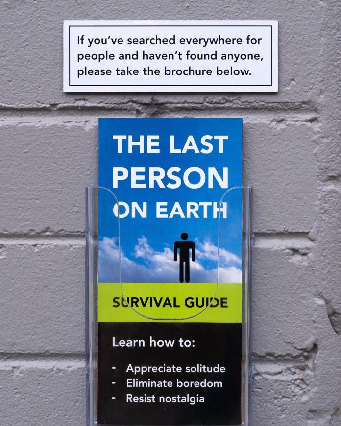 A brochure holder mounted on a brick wall contains a pamphlet titled "The Last Person on Earth Survival Guide." The pamphlet promises to teach readers how to "Appreciate solitude, Eliminate boredom, Resist nostalgia." A sign above it humorously reads: "If you’ve searched everywhere for people and haven’t found anyone, please take the brochure below." This street art by Michael Pederson in Sydney, Australia, wittily comments on isolation and urban loneliness.
