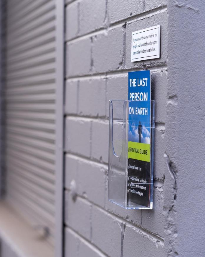 A side view of a brochure holder attached to a brick wall containing a pamphlet titled "The Last Person on Earth Survival Guide." The sign above the holder humorously reads, "If you’ve searched everywhere for people and haven’t found anyone, please take the brochure below." This artwork by Michael Pederson in Sydney, Australia, adds a satirical take on urban isolation.